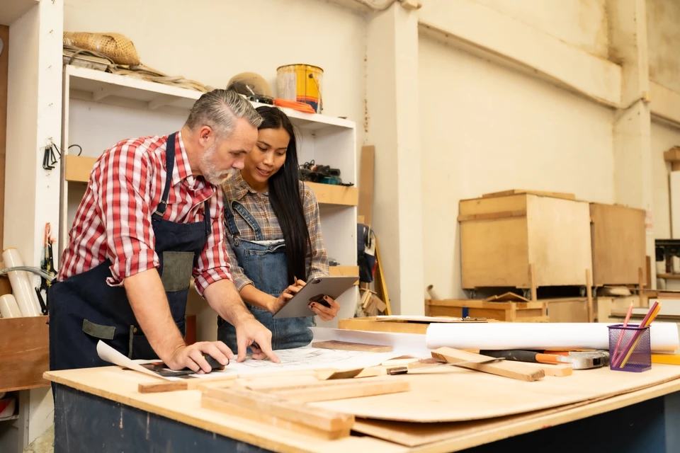 Atelier de menuiserie avec artisan expérimenté et apprentie consultant plans et tablette numérique