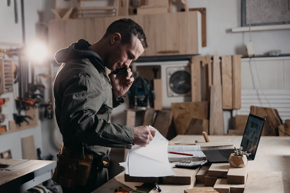 Atelier de menuiserie avec artisan en tablier de cuir consultant un carnet technique tout en inspectant des planches de bois brut
